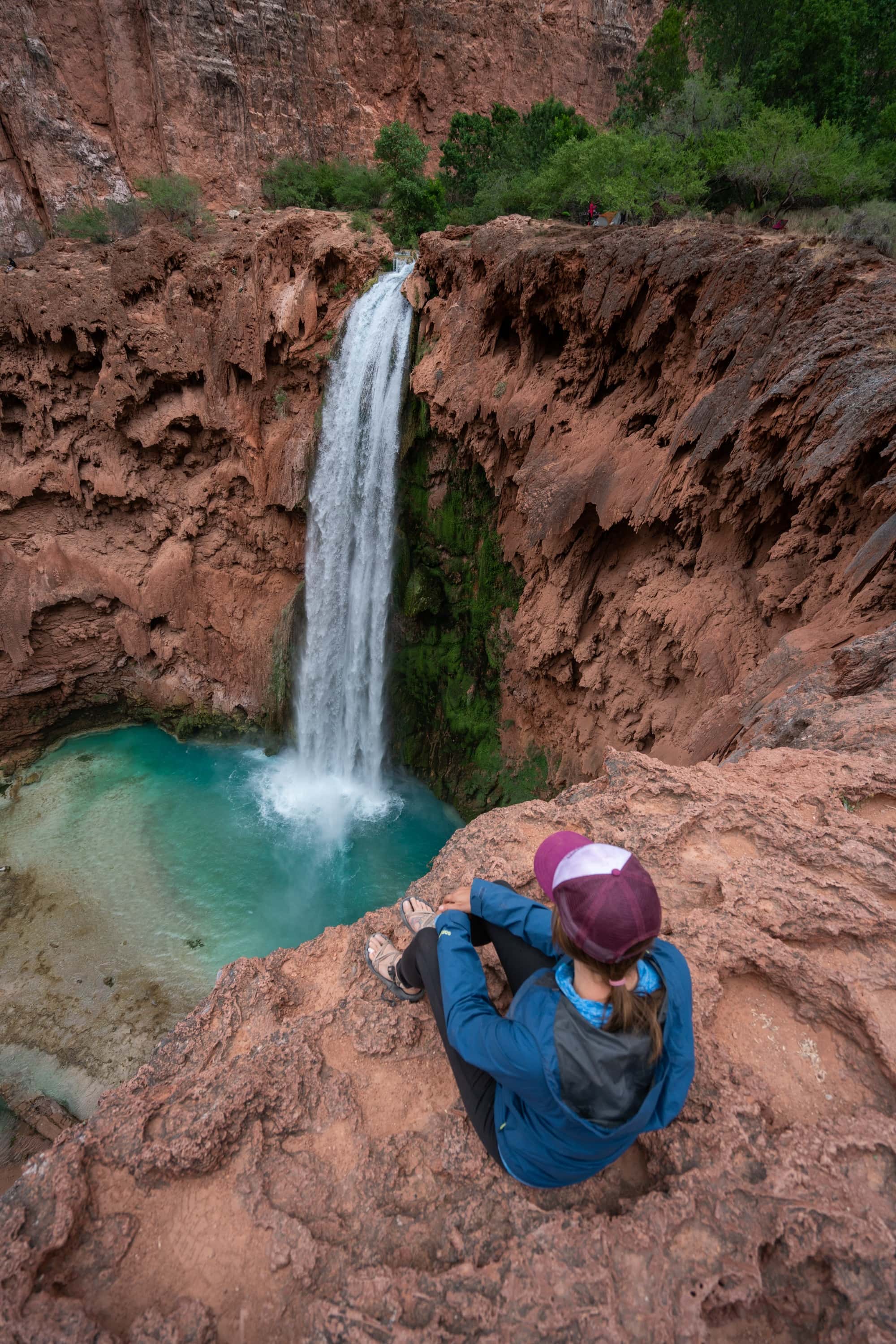 Woman sitting at the top of Mooney Falls in Havasu Canyon surrounded by red rock cliffs