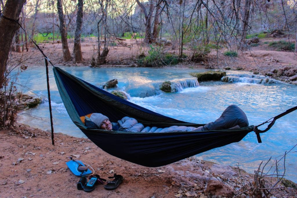 Woman lying in hammock next to river with blue water of Havasu Creek bundled in sleeping bag