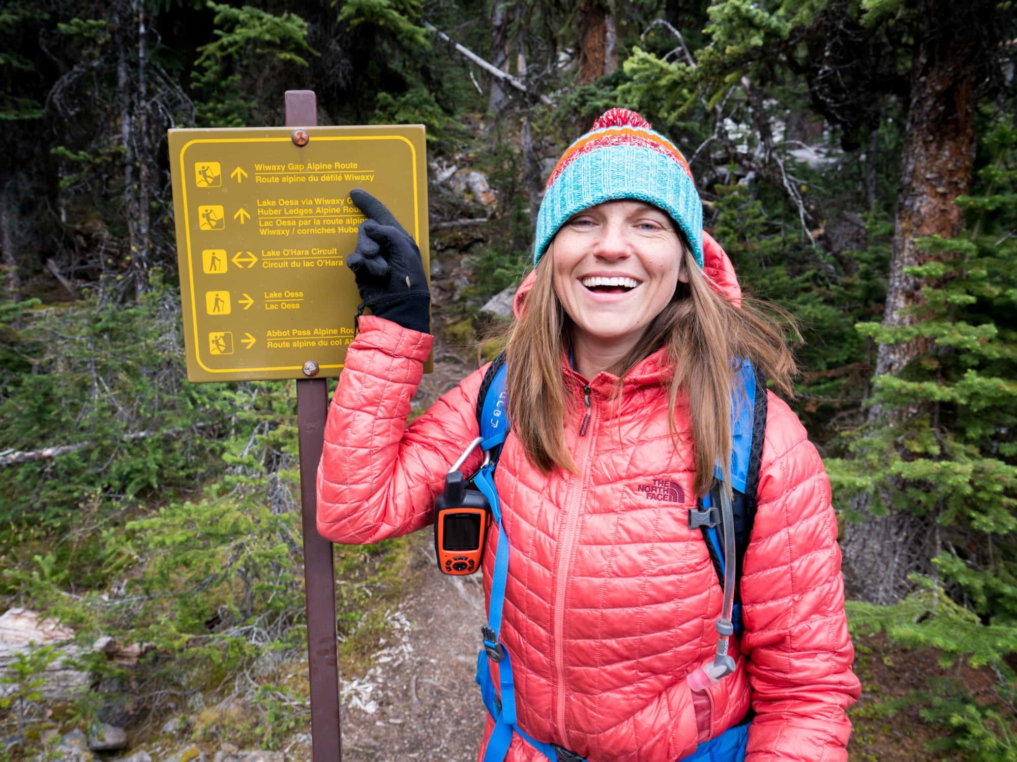 A woman points to a sign on a hiking trail. She is wearing a beanie, gloves, puffy jacket, and has the Garmin inReach Explorer clipped to her daypack