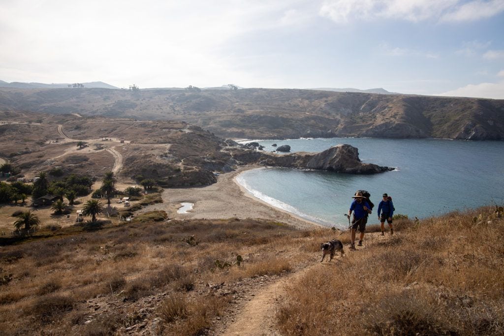 two backpackers hiking up trail on Trans-Catalina route on Catalina Island with ocean and harbor behind them