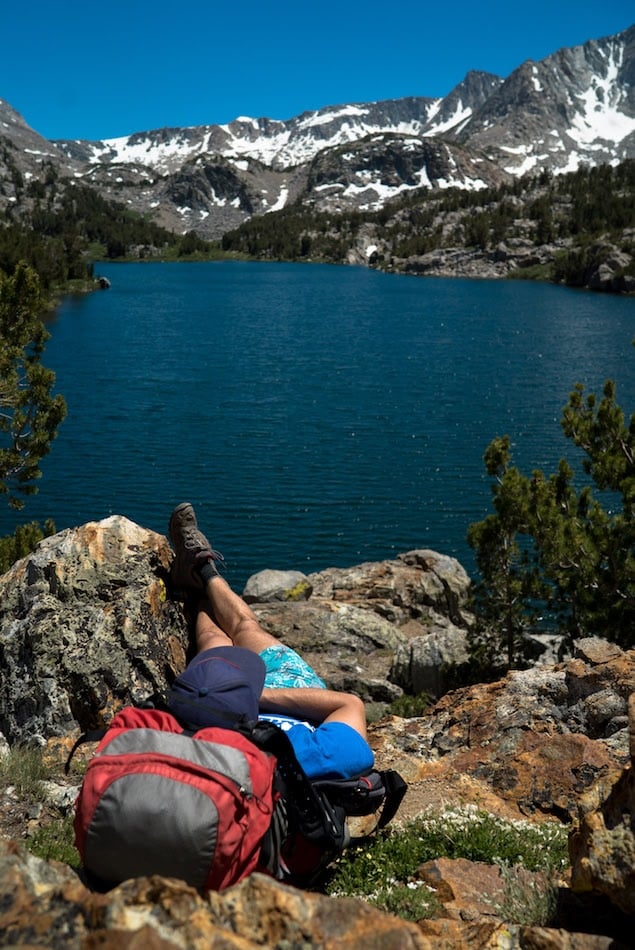 Person lying on rocks at alpine lake with snow patches on mountain peaks