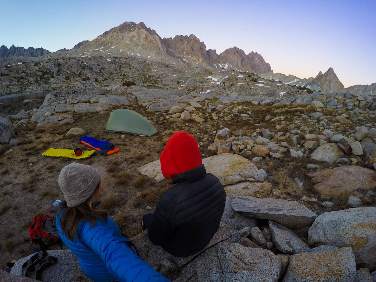 Two backpackers bundled up in warm clothes sitting at remote campsite in Dusy Basin looking out over mountain peals and rock field