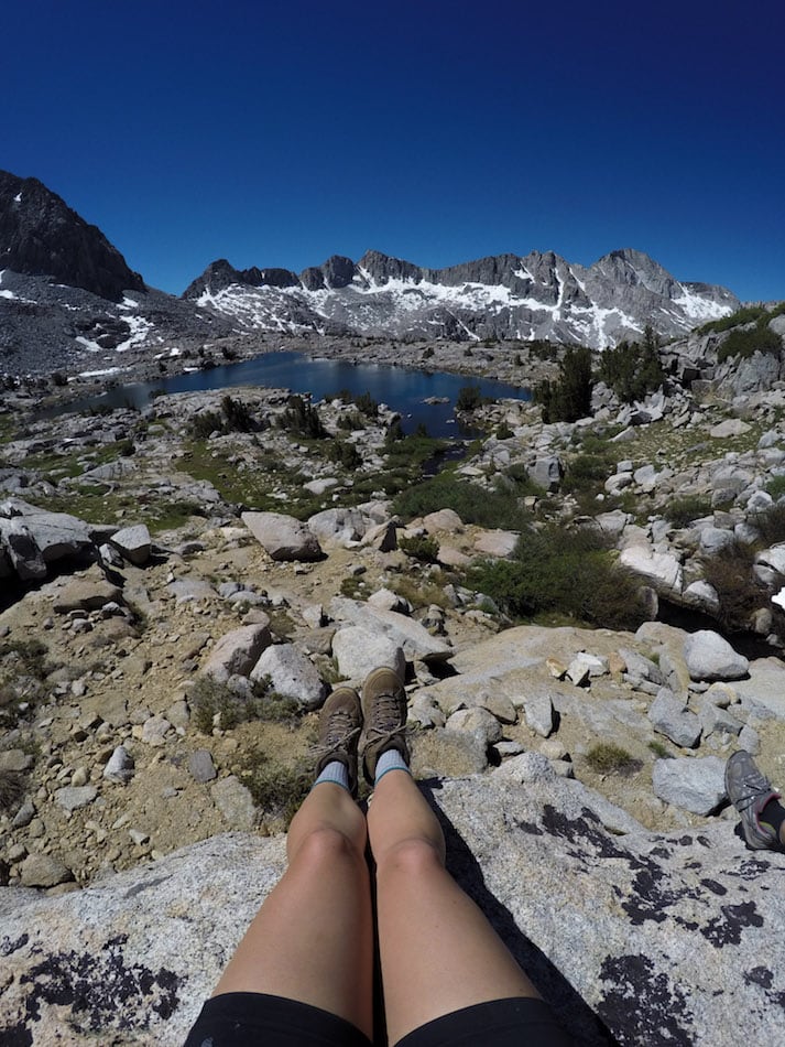 Photo out over women's legs sitting on rock overlook onto scenic alpine landscape with lakes, mountains, and high alpine trees