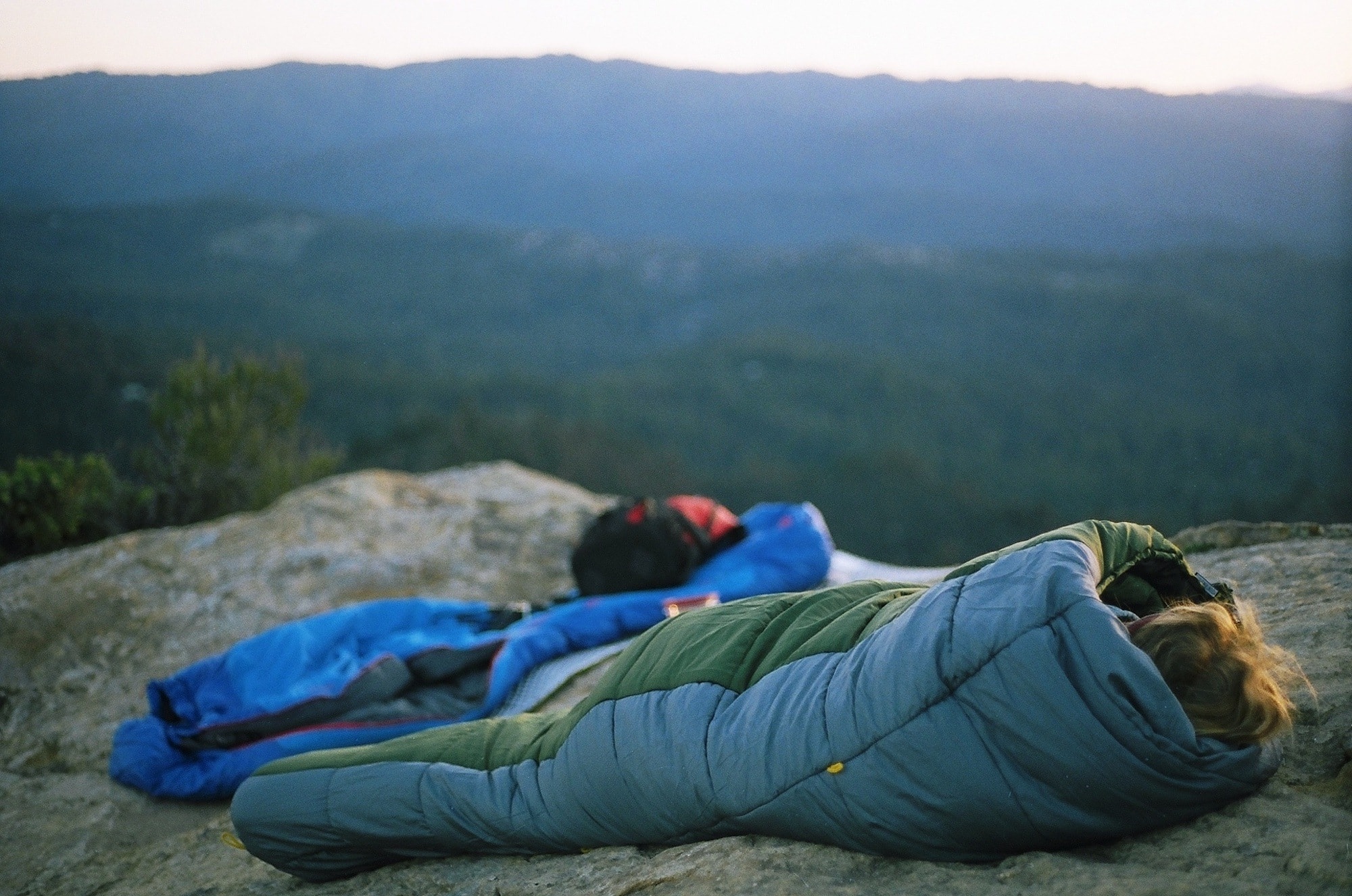 a woman cowboy camping on a rocky summit with a grey and green sleeping bag