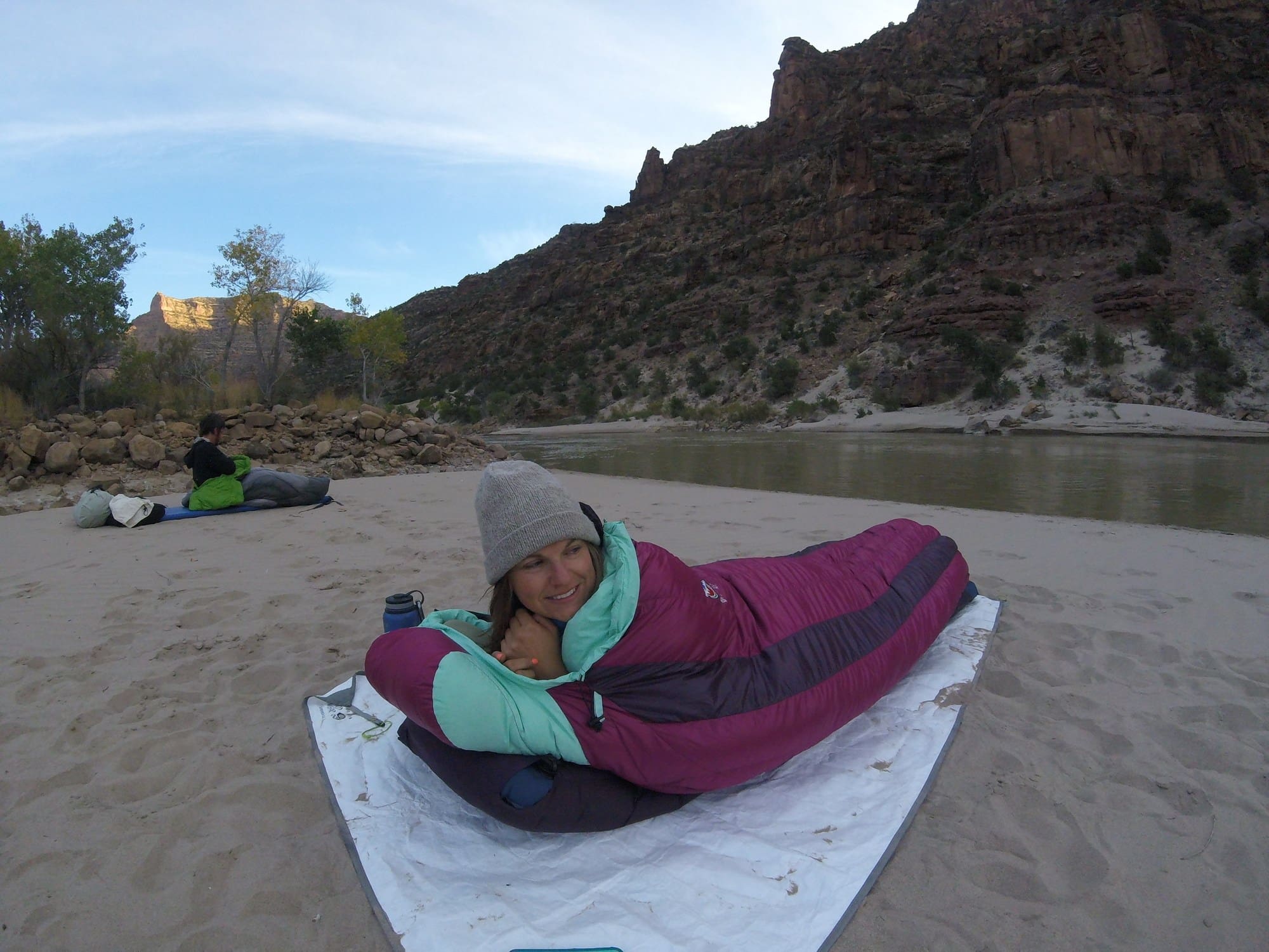 woman cowboy camping in a purple sleeping bag on the sand next to a river in a small canyon