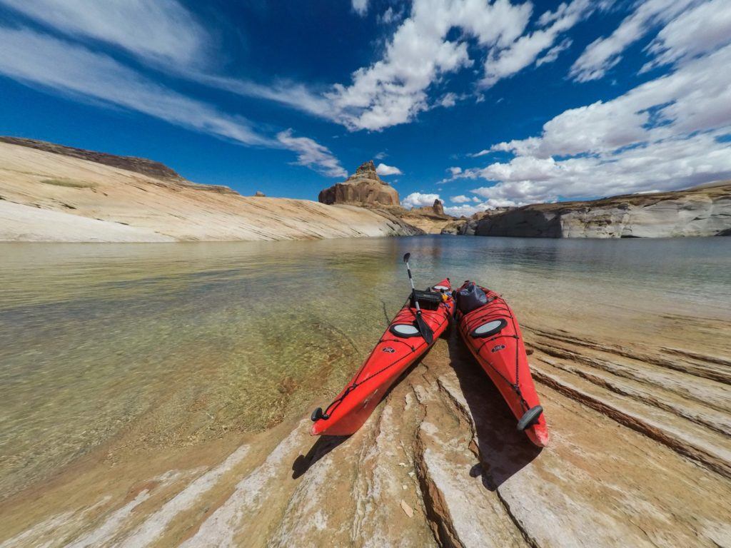 Two kayaks are beached on a rock ledge shore overlooking the calm, blue-green waters of Lake Powell. The sun is shining brightly, illuminating the surrounding red rock cliffs and casting a reflection on the glassy surface of the lake. In the distance, sandstone bluffs rise up from the water, creating a serene and picturesque landscape. The kayaks are brightly colored, with paddles resting on the sand next to them, inviting the viewer to imagine an adventure on the tranquil waters of the lake.