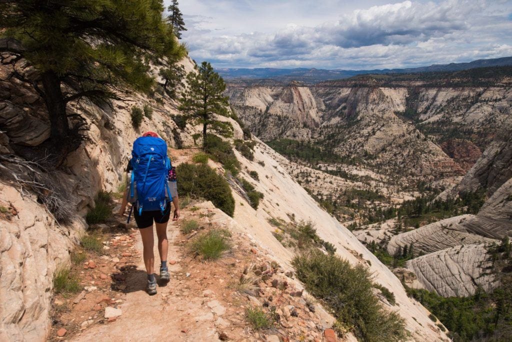 Hiker walking down narrow section of trail with rock wall on left side and steep rock slab drop off on right