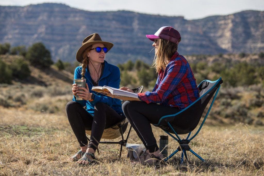 Two women sitting in camping chairs facing each other and chatting with tall bluffs in background