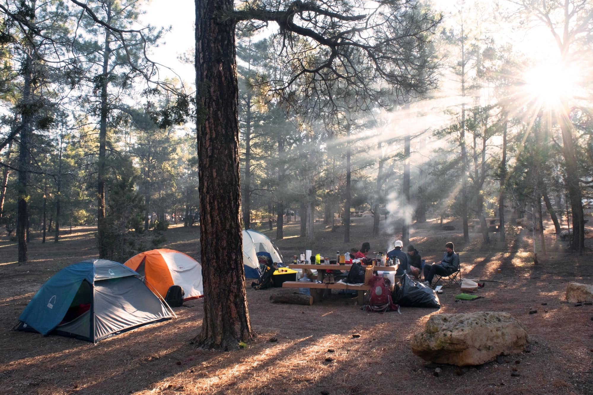 Three tents at a campsite with trees and a group of people sitting around a fire in camp chairs with items on a camp table nearby.