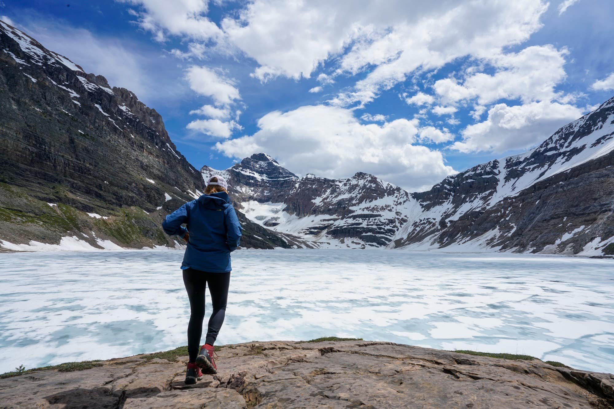 Hiker standing on flat rock in front of iced-over Lake McArthur in Yoho National Park, British Columbia. Snow covered peaks are in the background.