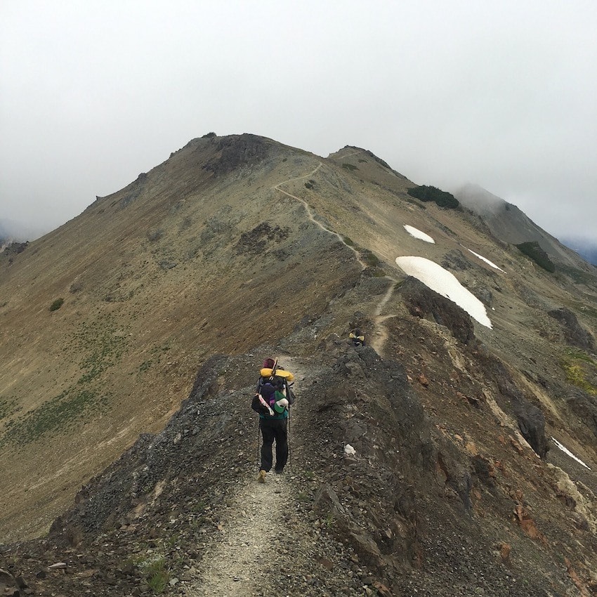 Pacific Crest Trail thru-hiker carrying backpack and gear along narrow ridge trail 