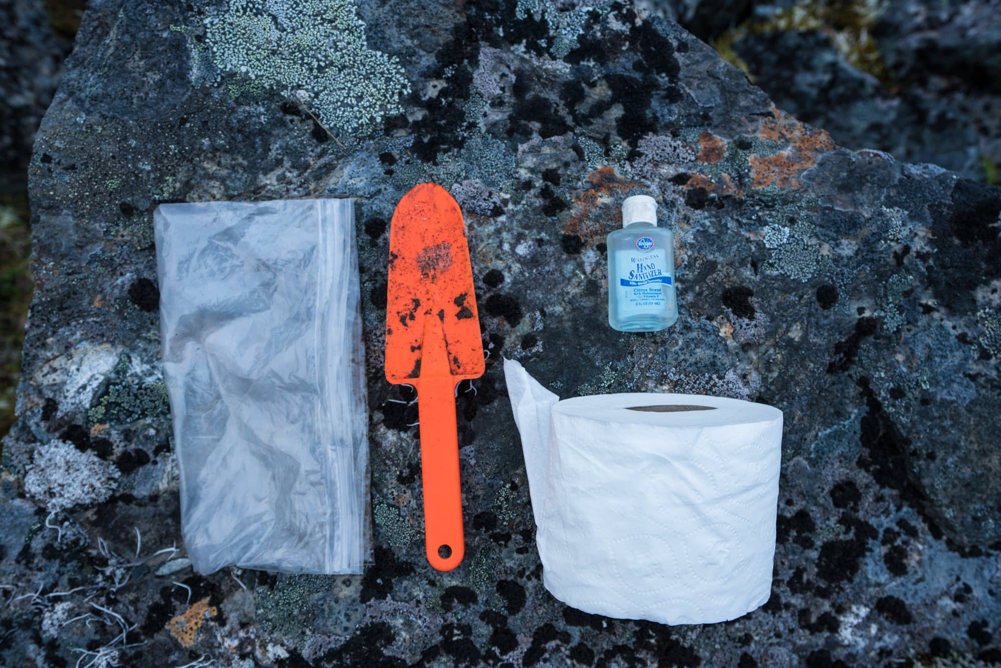 A plastic bag, orange trowel, bottle of hand sanitizer, and toilet paper roll spread out on a rock outside