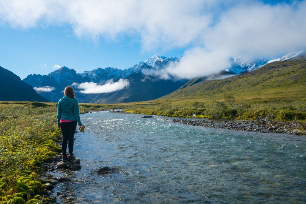 Kristen wearing standing next to river in Alaska looking out snow-capped mountain range