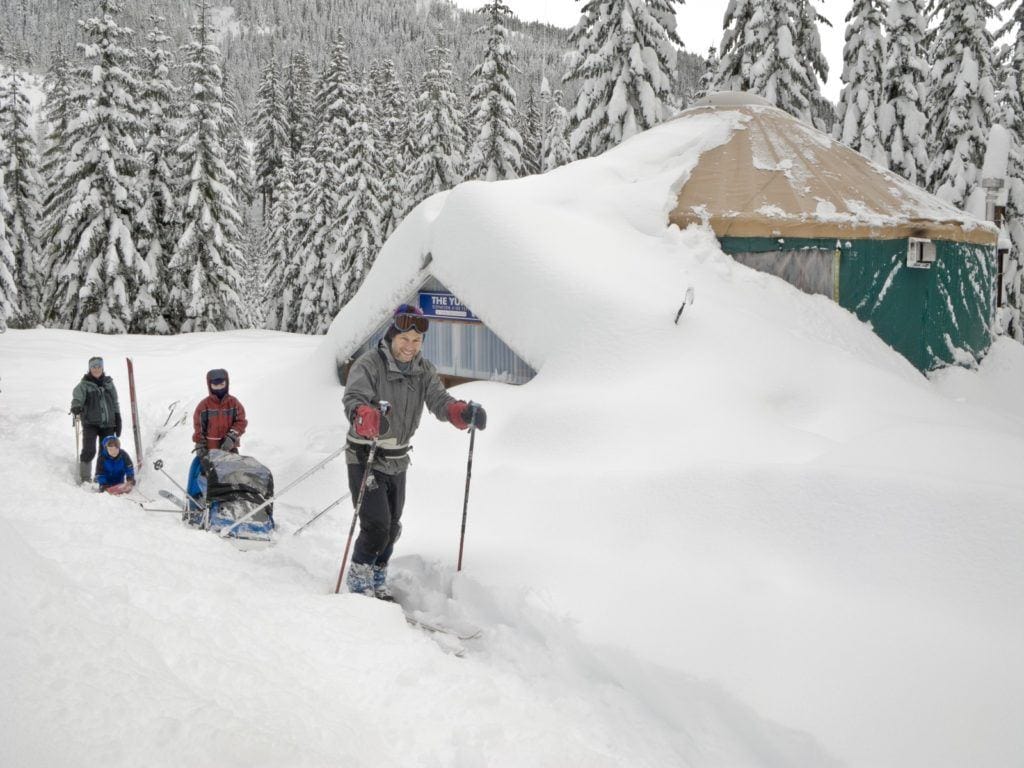 Man on skies pulling sled loaded with gear on trail next to snow-covered backcountry yurt and followed by family of three