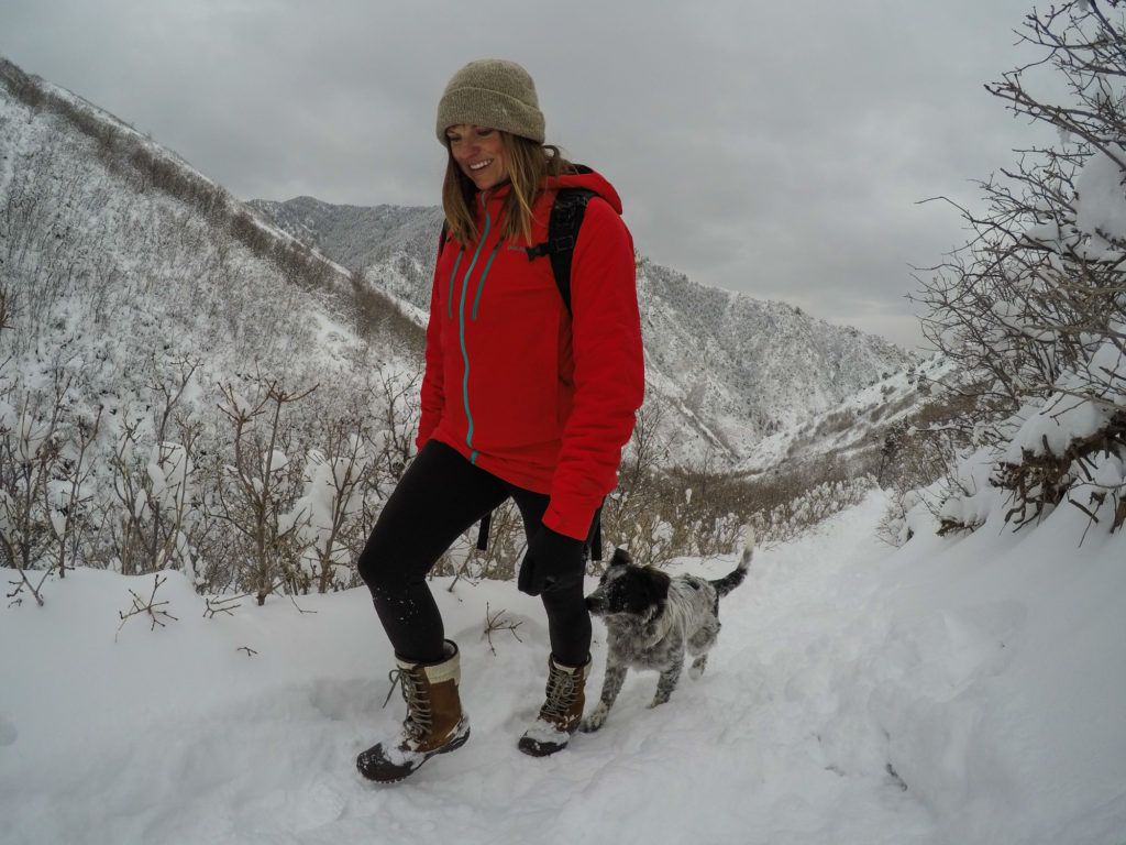 Woman on snowy hiking trail in mountains with puppy following behind