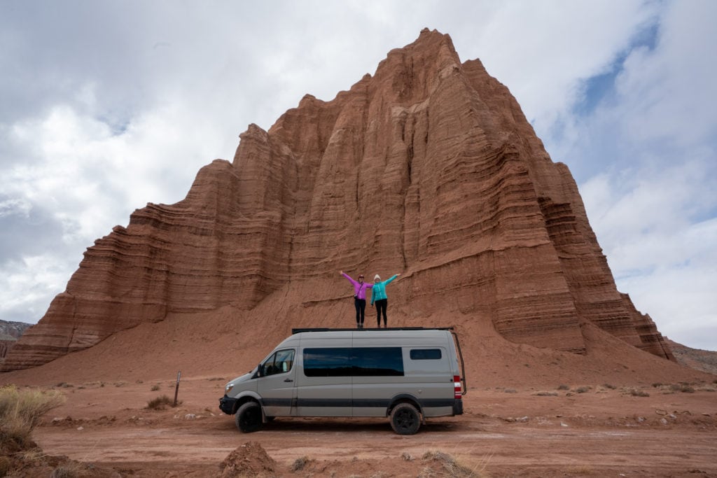 Two people standing on the top of Sprinter van in front of massive red rock monolith in Capitol Reef National Park