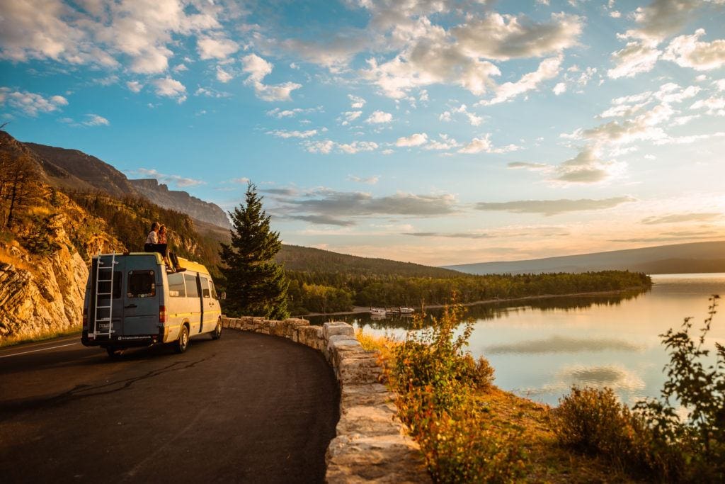 Two people sit on the roof of a camper van at a pull out near a lake