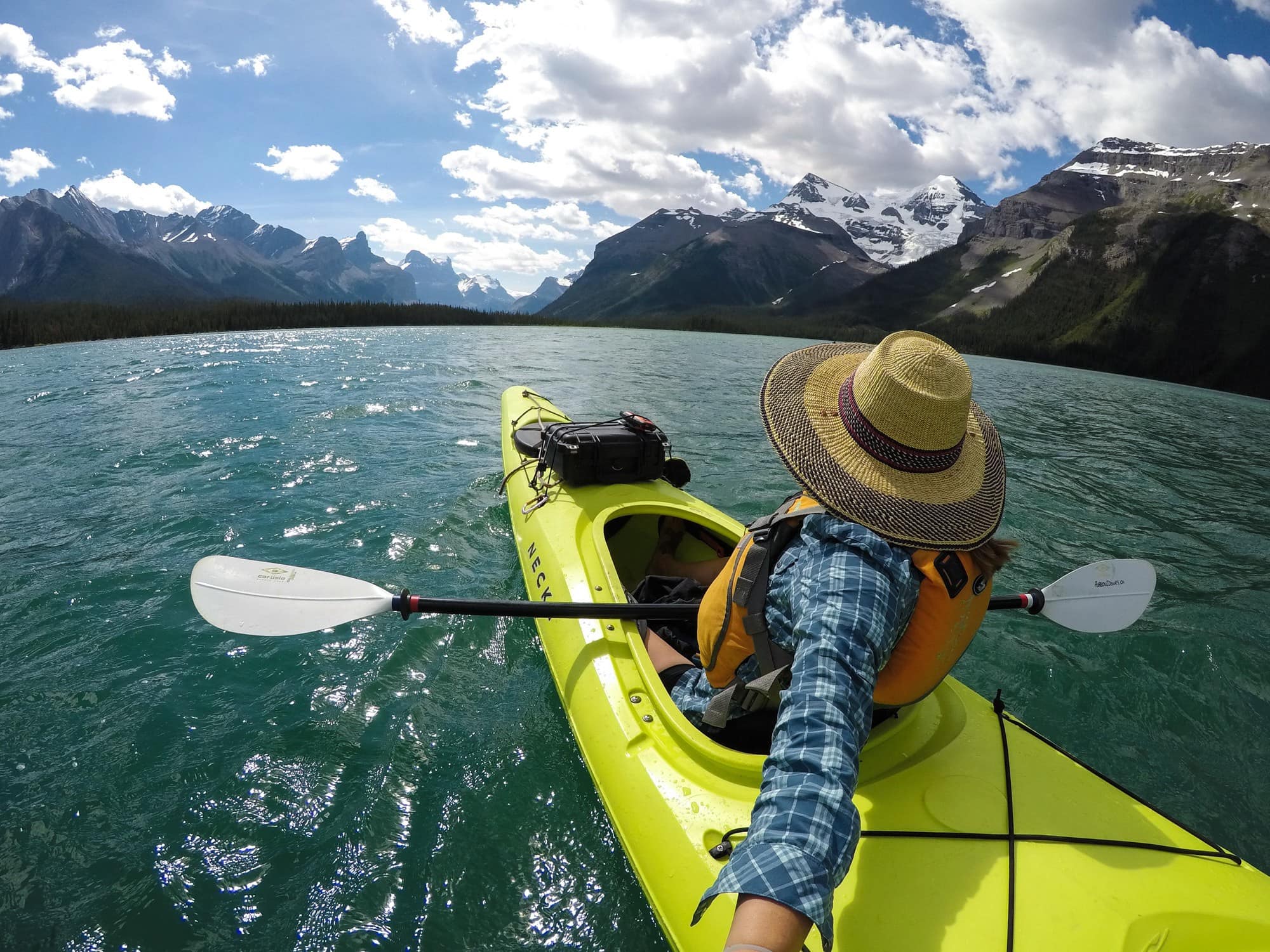 A woman takes a selfie from behind while kayaking. She's in a yellow sit-in kayak and wearing a sun hat.