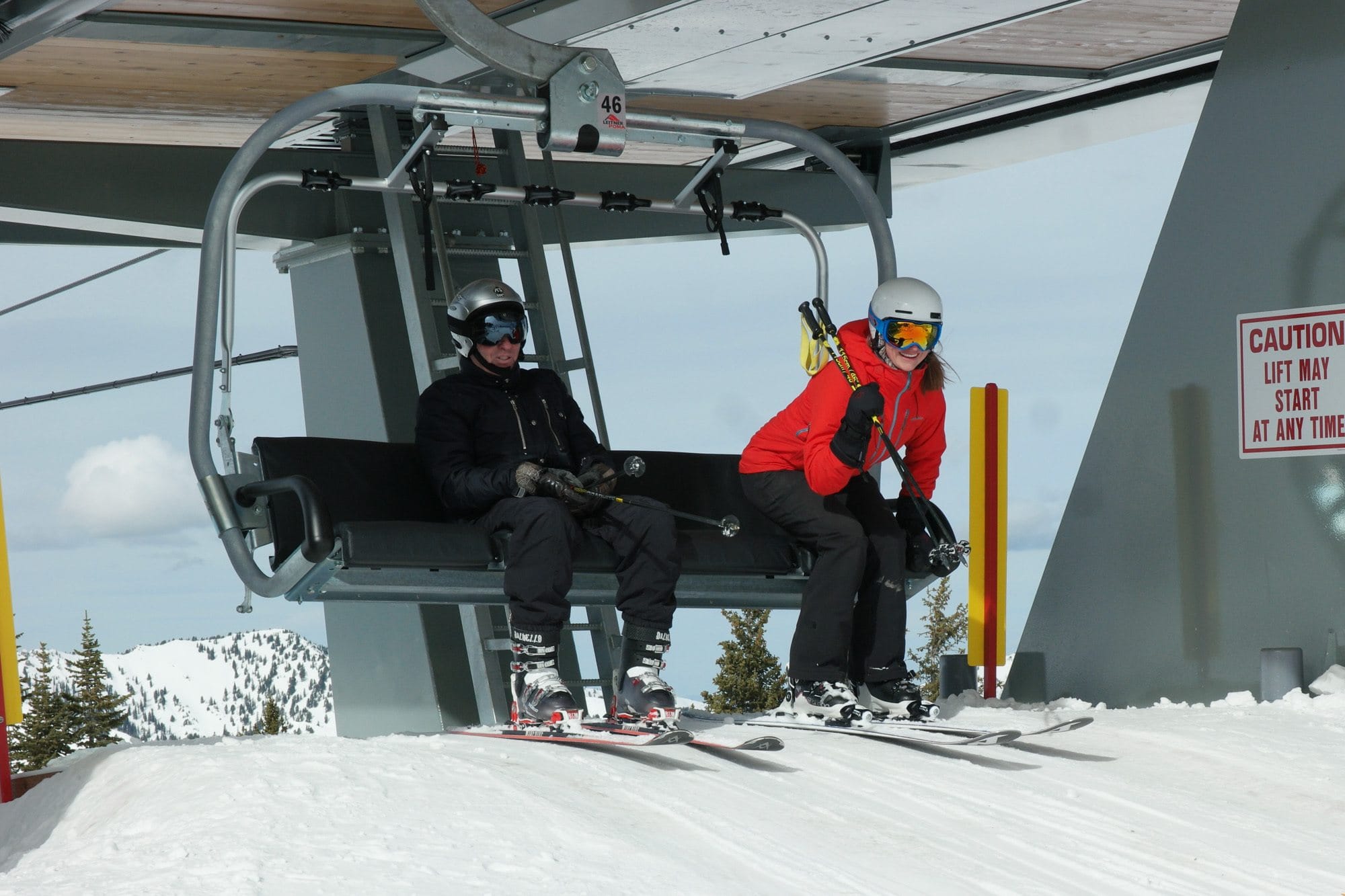 A man and woman sitting on a chairlift wearing skis and snow gear