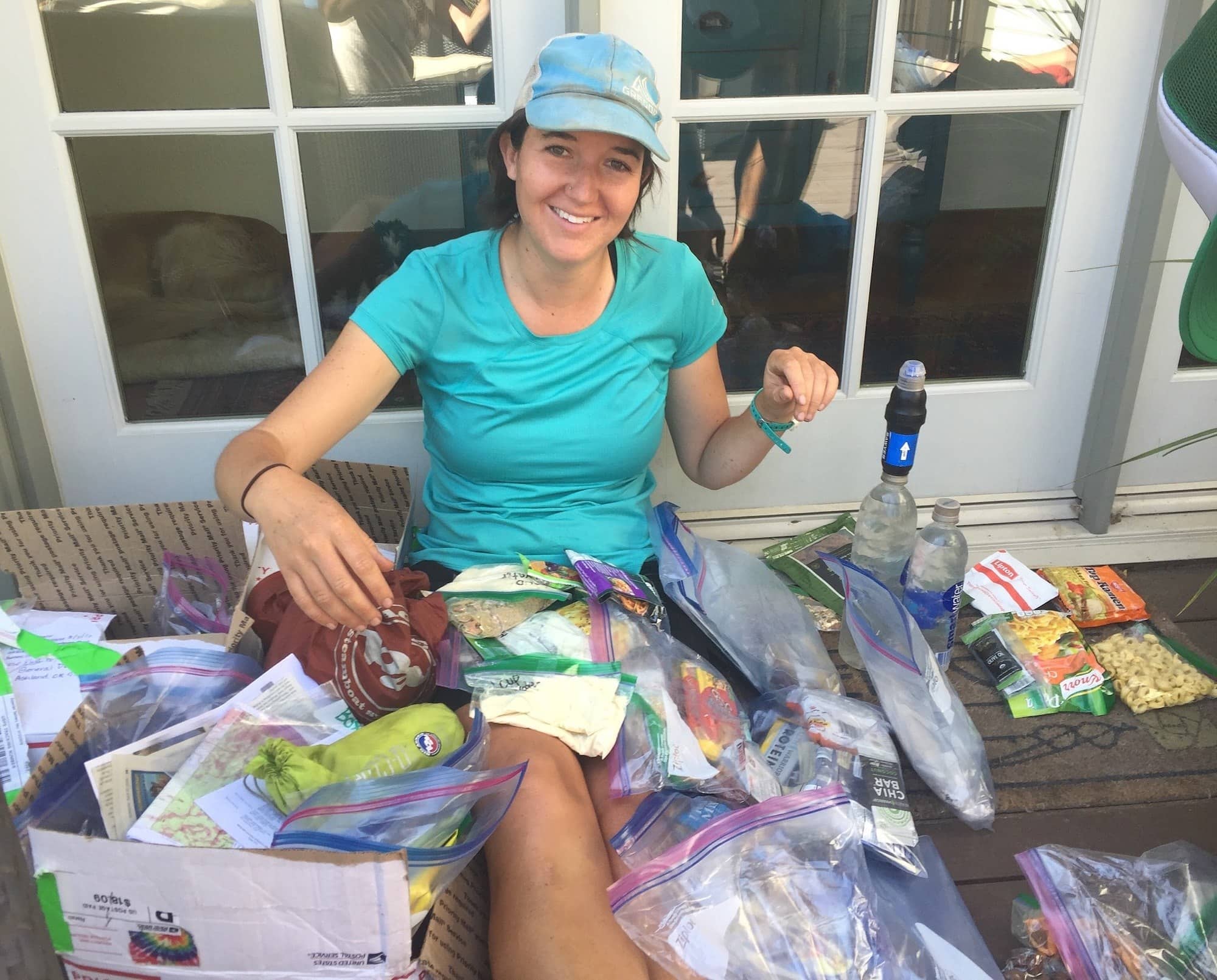 woman sitting on the ground smiling with piles of PCT resupply food items and a USPS mailing box