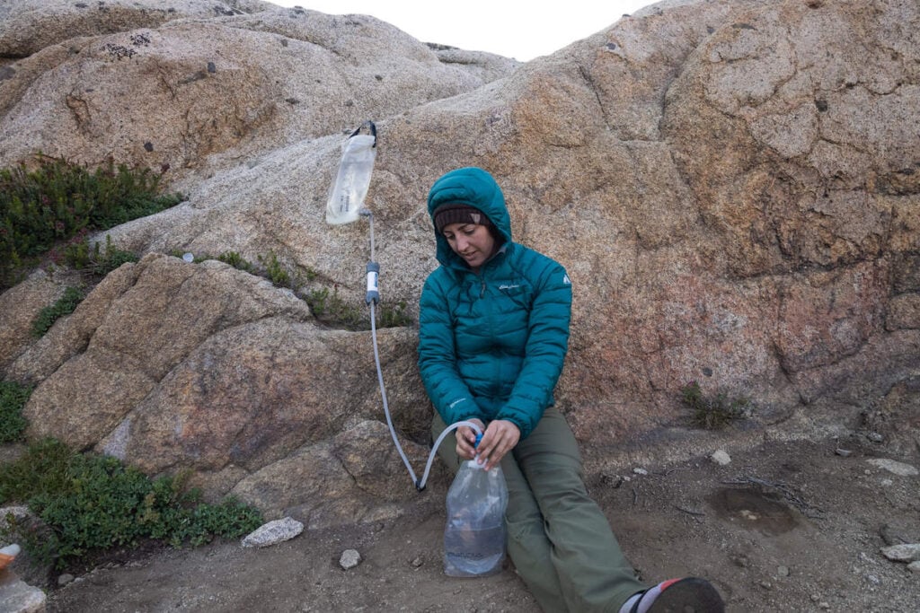 Woman sitting on ground in front of large rock filtering water through a Platypus GravityWorks backpacking water filter