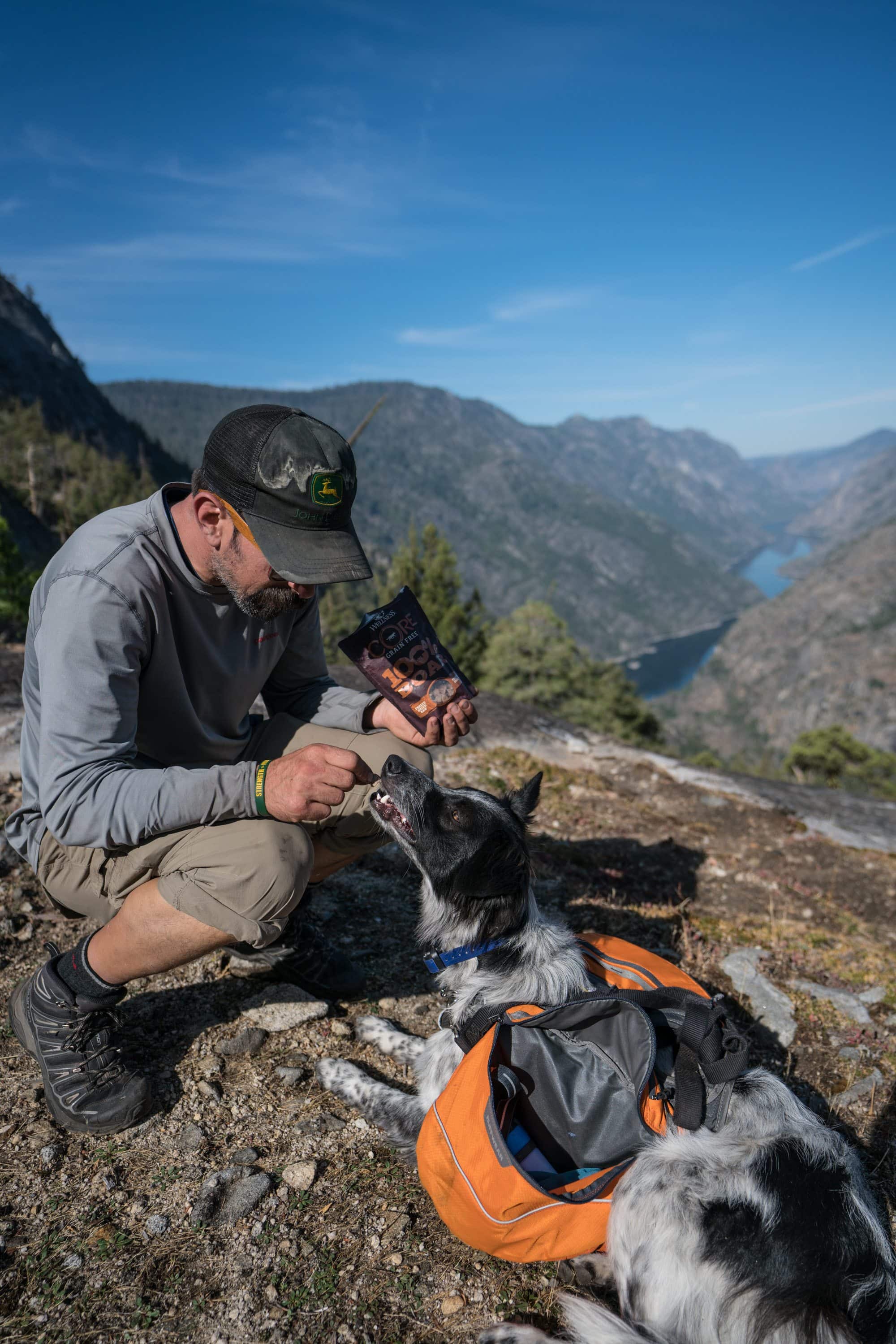 Man feeding dog treats while hiking. Dog is wearing a dog backpack