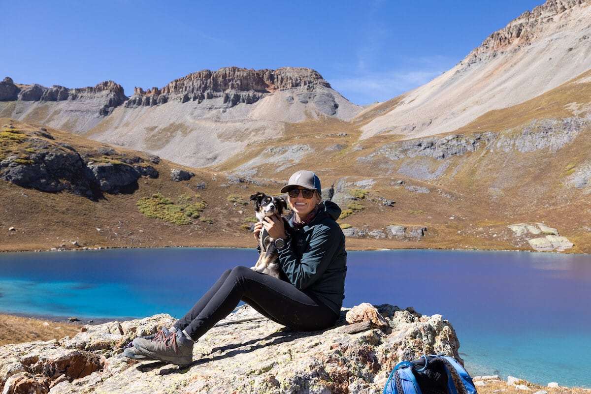 A woman sitting holding a puppy on a hike near a blue lake wearing PrAna Transform leggings