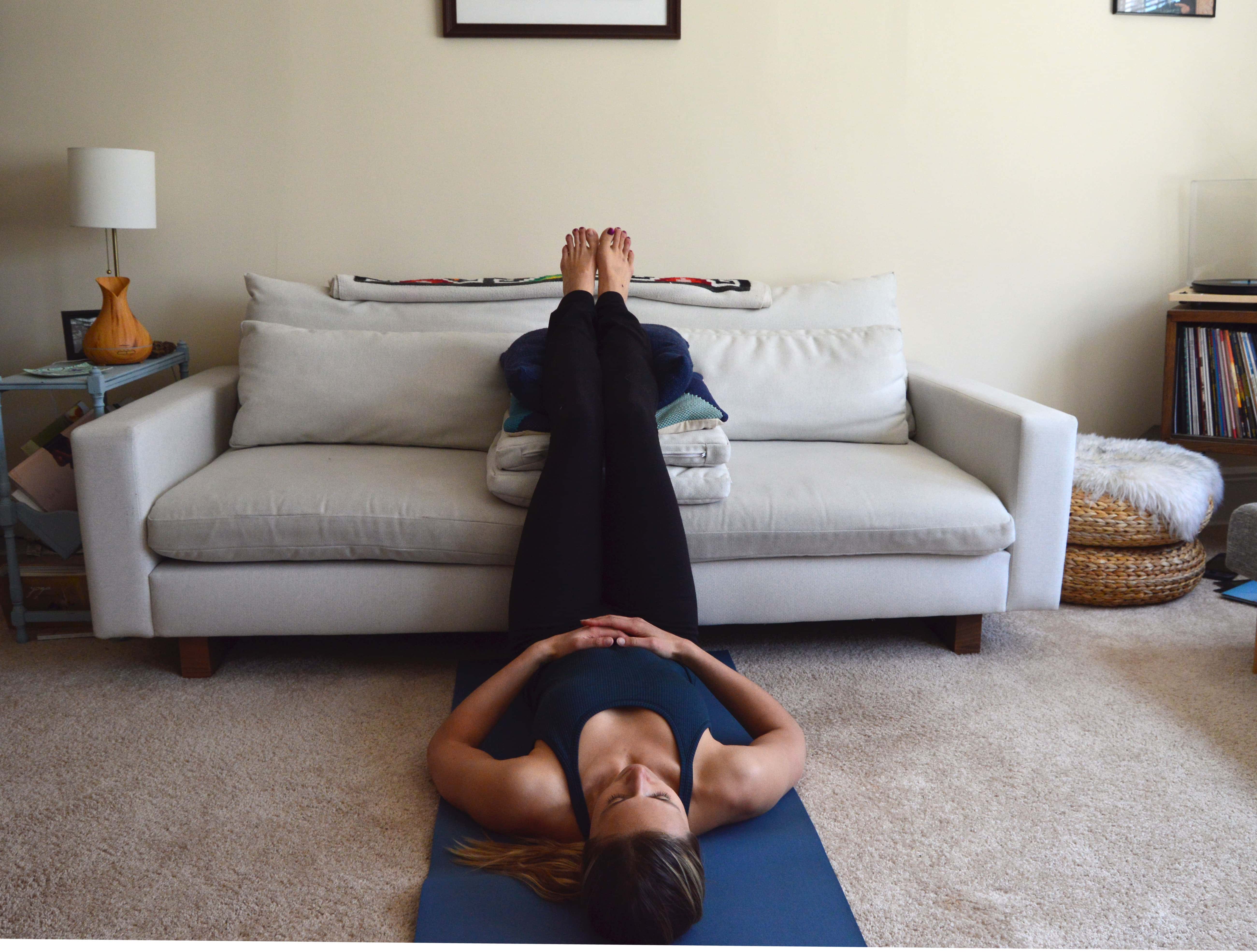 A woman lays on a yoga mat on the floor with her legs raised on the couch