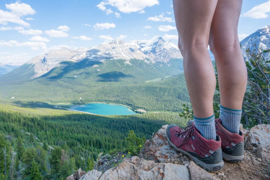 A woman wears a pair of hiking boots  on a hiking trail. There is a lake and mountains in the distance.