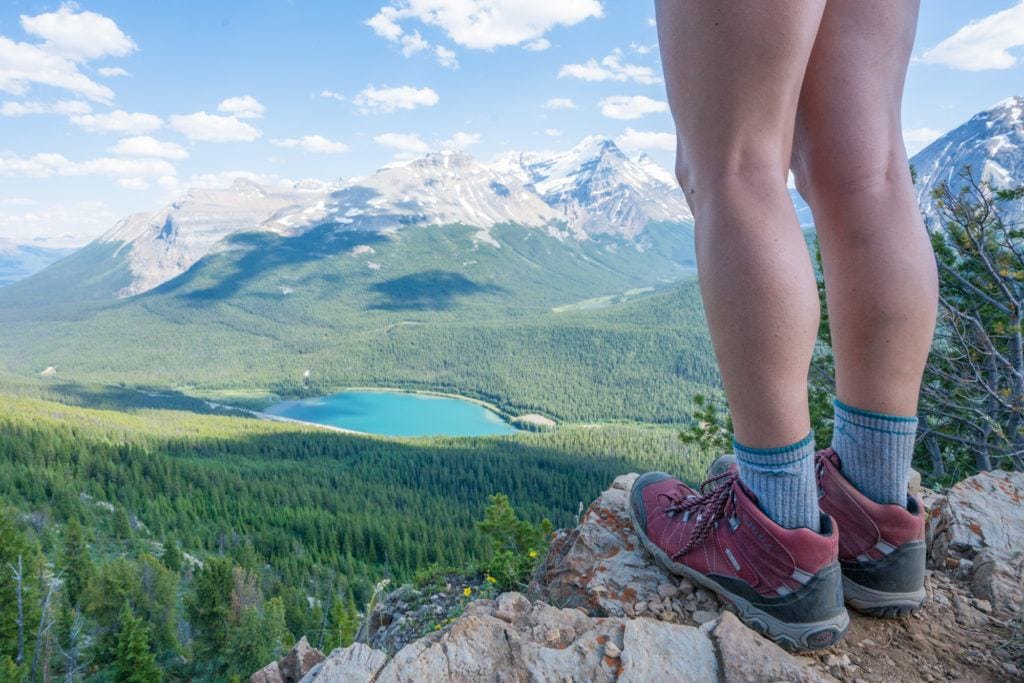 A woman stands on a summit looking over a blue lake and lots of alpine trees. She's wearing red Oboz hiking boots