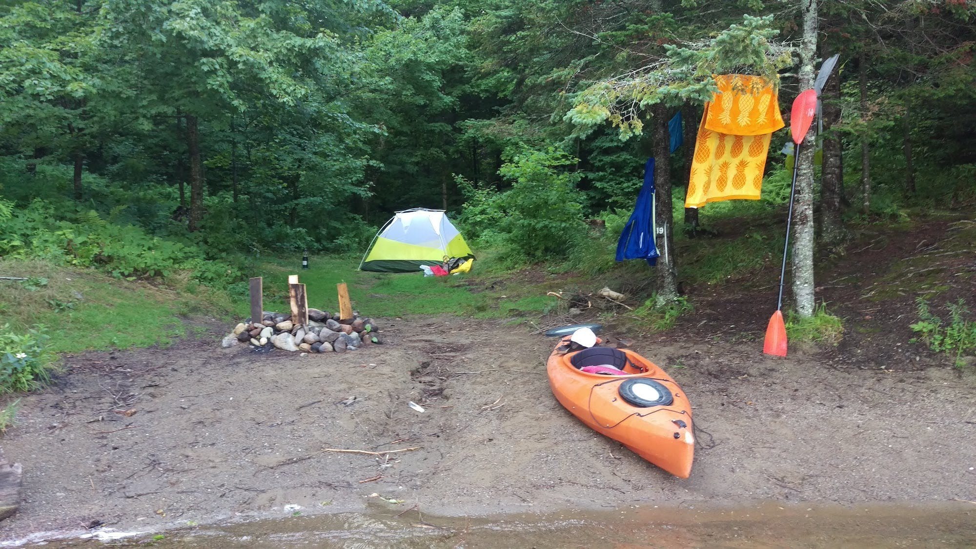 Kayak pulled up on lake short with fire pit, tent, and camp gear laid out around camp