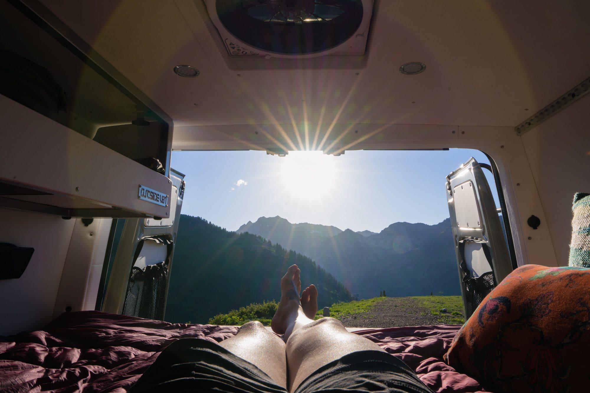 A woman lays on her bed in the back of her Sprinter van with the back doors open. The sun is shining. She is laying on a Rumpl blanket.