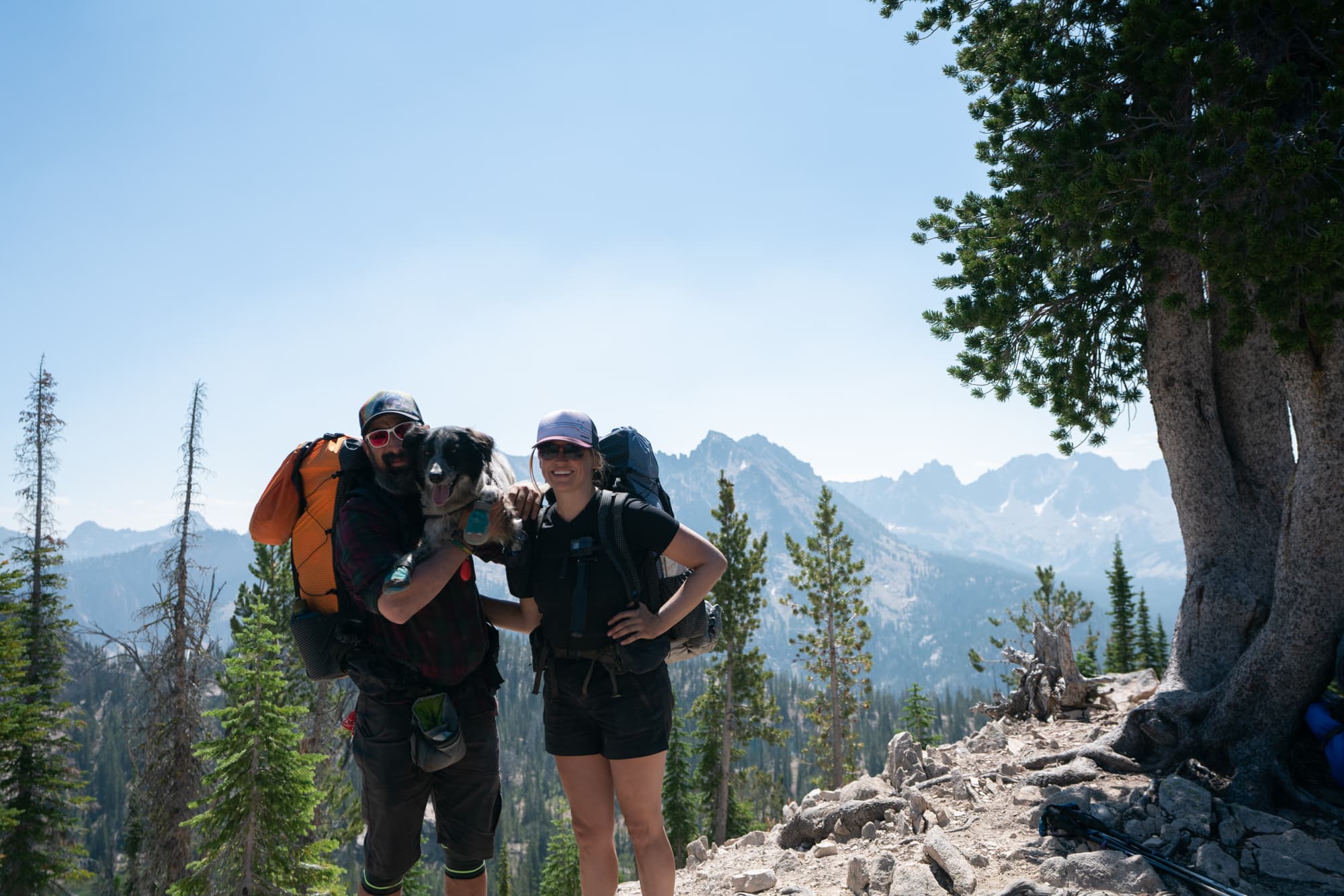 Two backpackers and their dog standing for a photo on Baron Lake trail in Idaho with mountain range in the distance