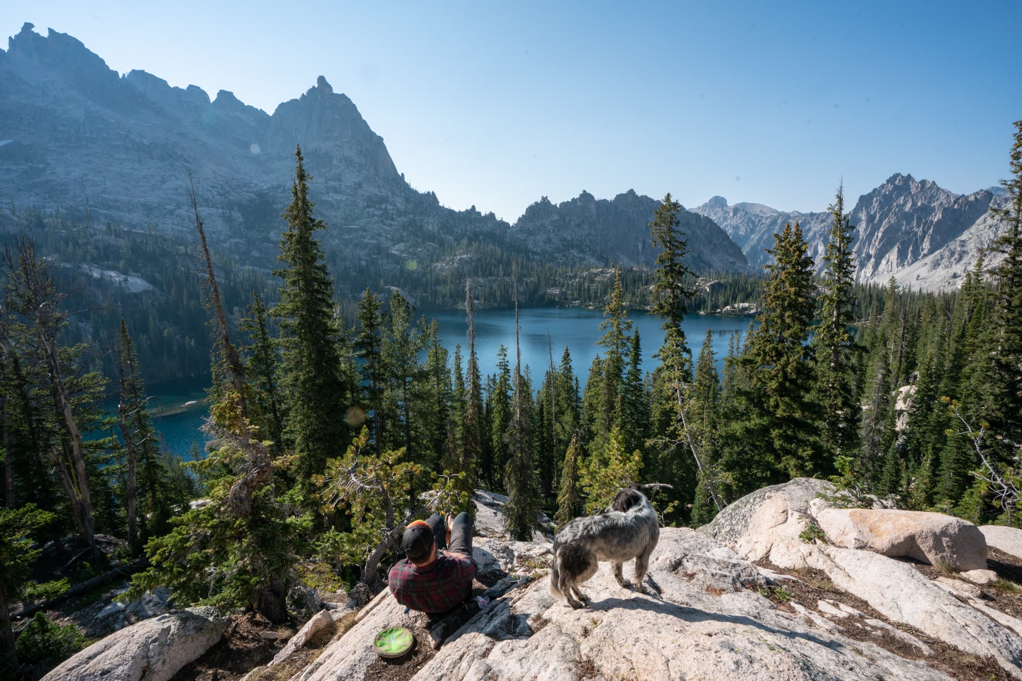 Man sitting on rock slab overlook with his dog. Baron Lake and Sawtooth Mountain landscape in front of him