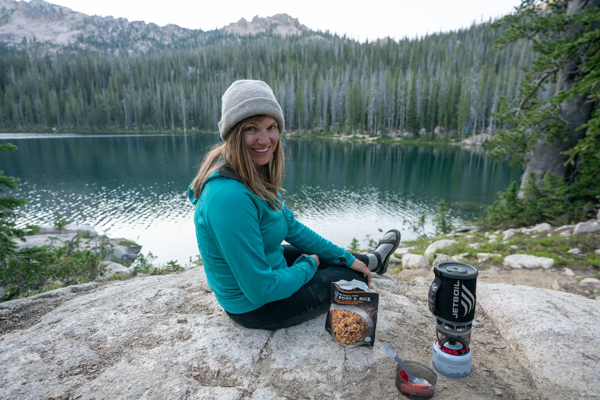 Woman sitting on large rock slab overlooking an alpine lake. She is boiling water in a camp stove for a dehydrated meal