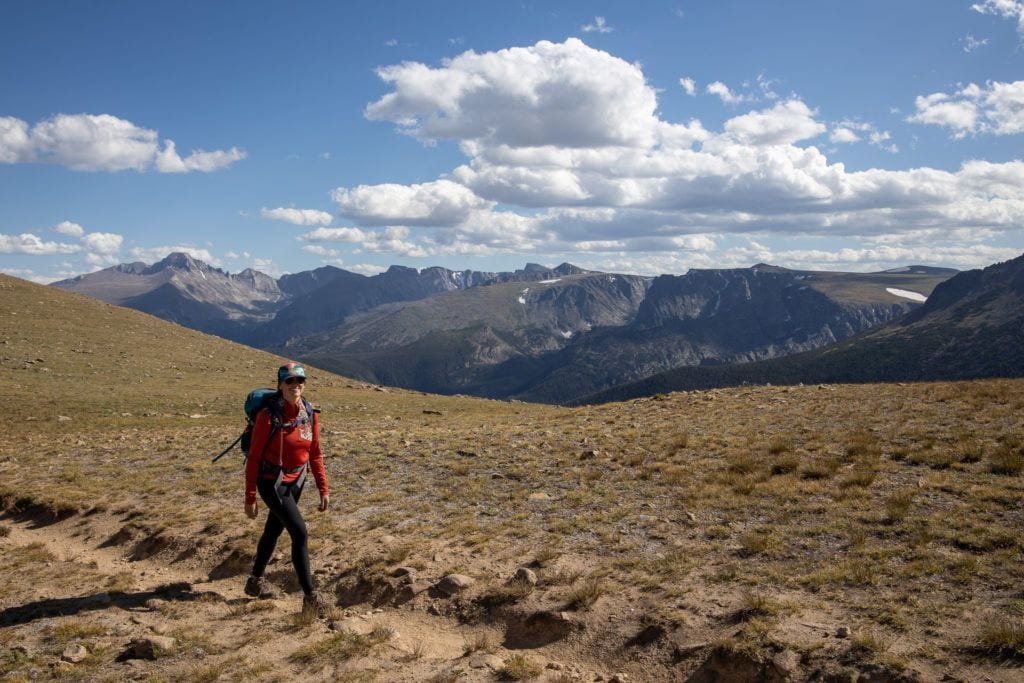 A woman smiles while hiking on a trail. There are mountains in the background.