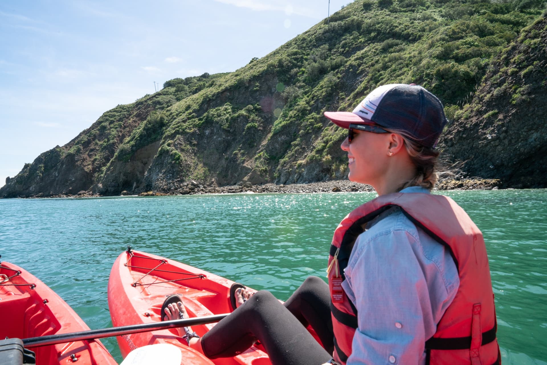 A woman smiles sitting in a red sit-on-top kayak. She is wearing a PFD