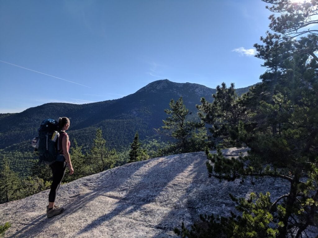 Woman standing on rock slab carrying backpacking pack looking up at mountain ridgeline