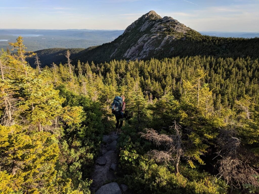 Woman hiking down trail through stunted pine trees carrying a backpacking pack with mountain views in the distance