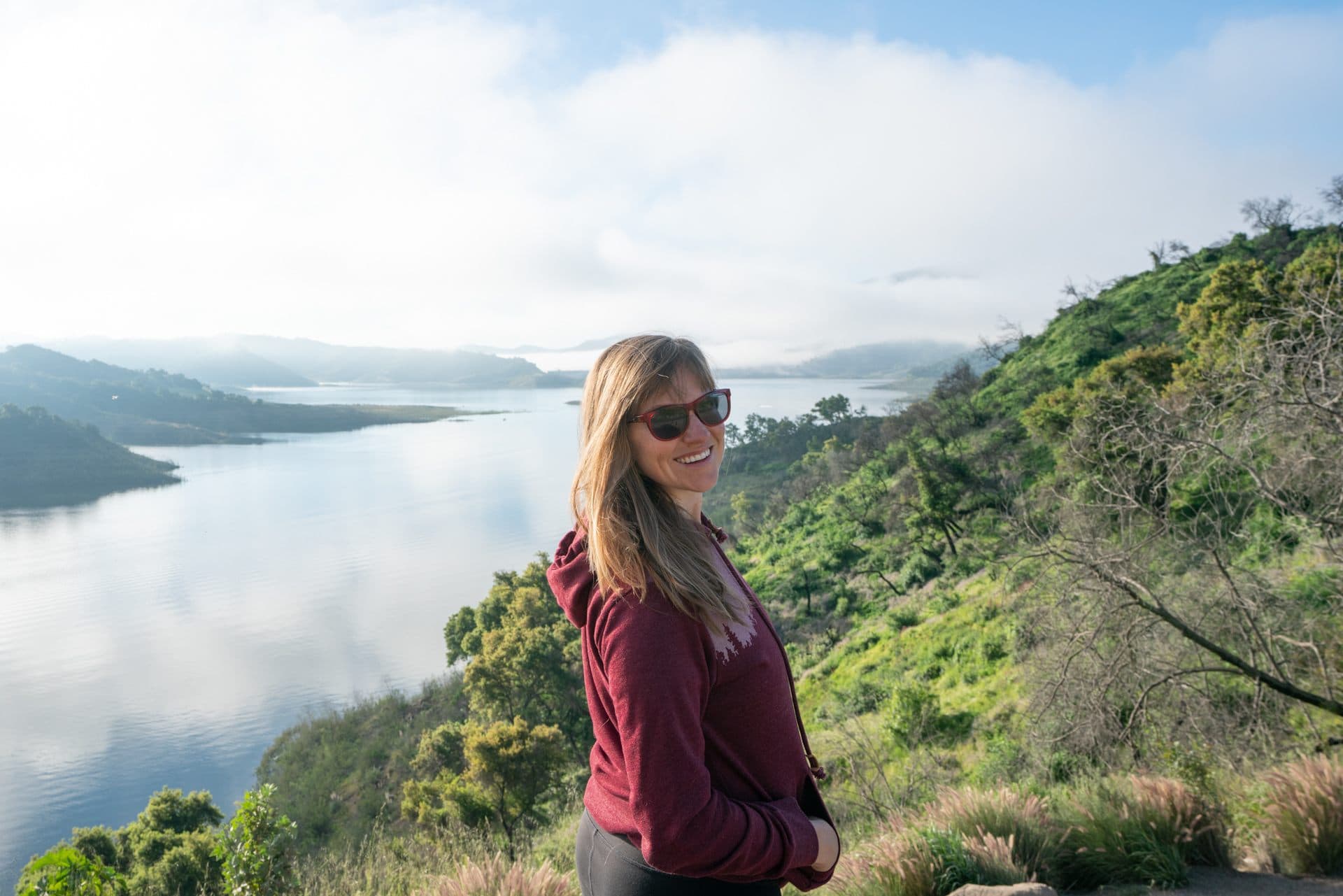 Woman wearing a maroon tentree hoodie while hiking in Central California with the ocean and green hills in the background