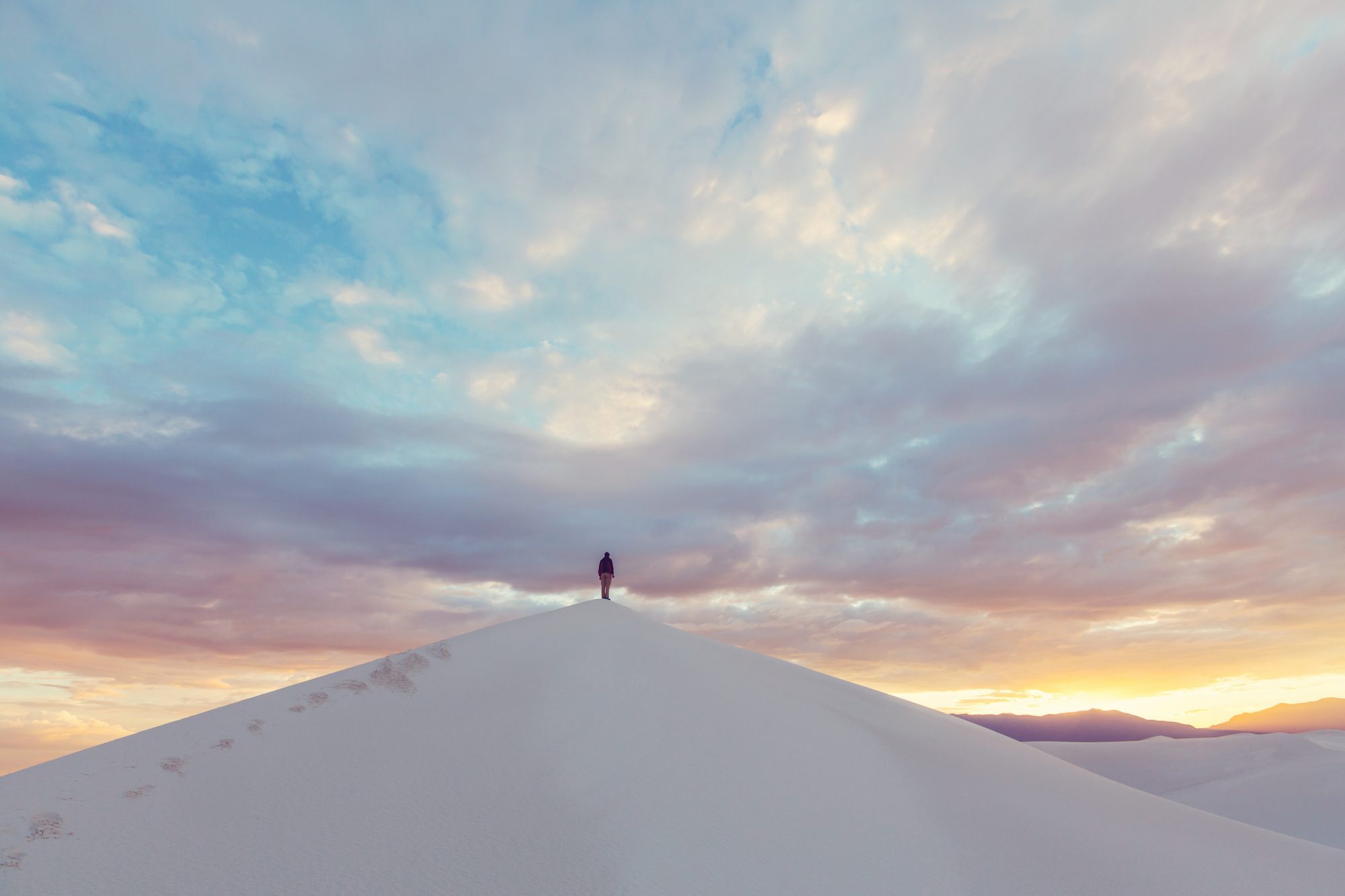 Person standing on top of large sand dune in White Sands National Park with a colorful sunset sky overhead