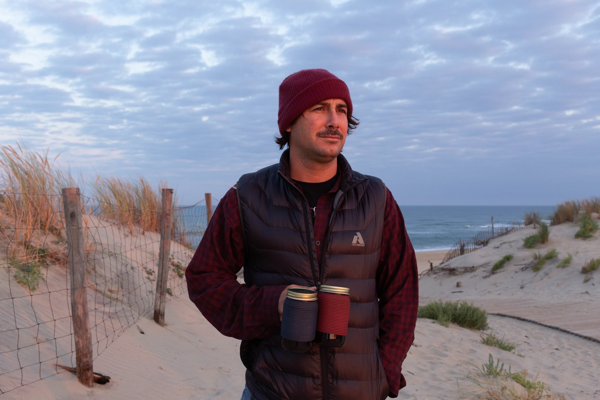 Man carrying two insulated mugs on beach at sunset wearing warm layers