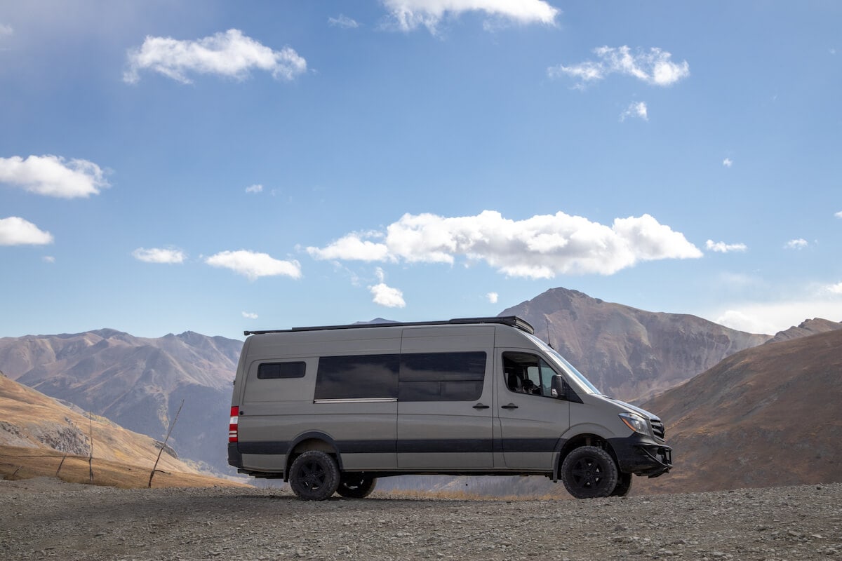 A sprinter van with mountains behind it along with a blue sky and clouds.