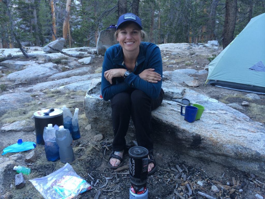 A woman smiles making food on her JetBoil on the JMT