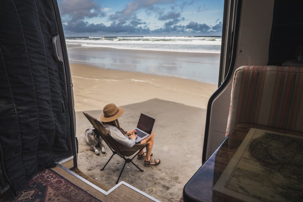 A woman sits in a camp chair outside of her Sprinter van on beach. She is working on her laptop and a dog is laying next to her.