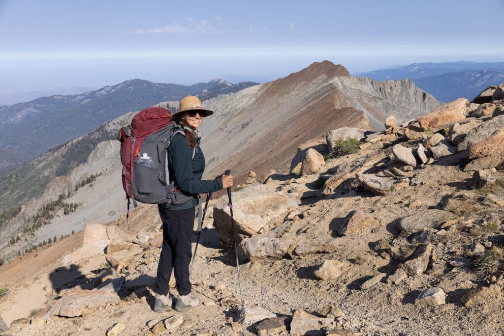 A woman smiles wearing a red backpacking pack on top of a rocky ridge wearing Mountain Hardwear Dynama Ankle Pants in Sequoia National Park