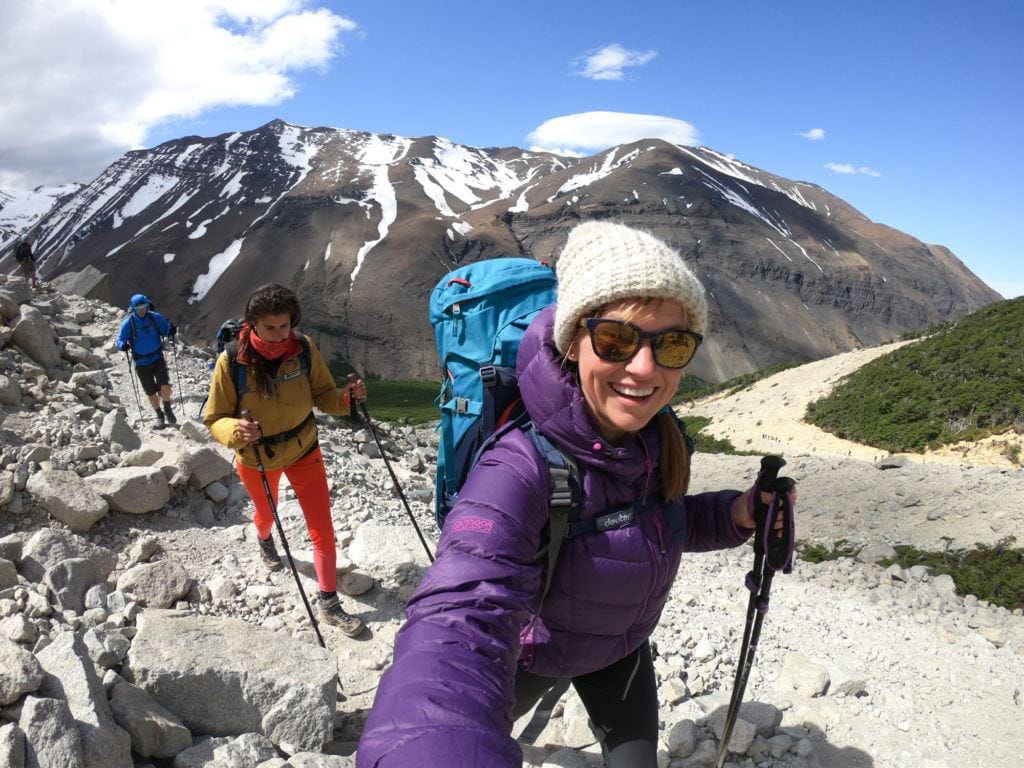 Kristen taking a selfie on hiking trail with two other hikers in back and snowy mountain in background