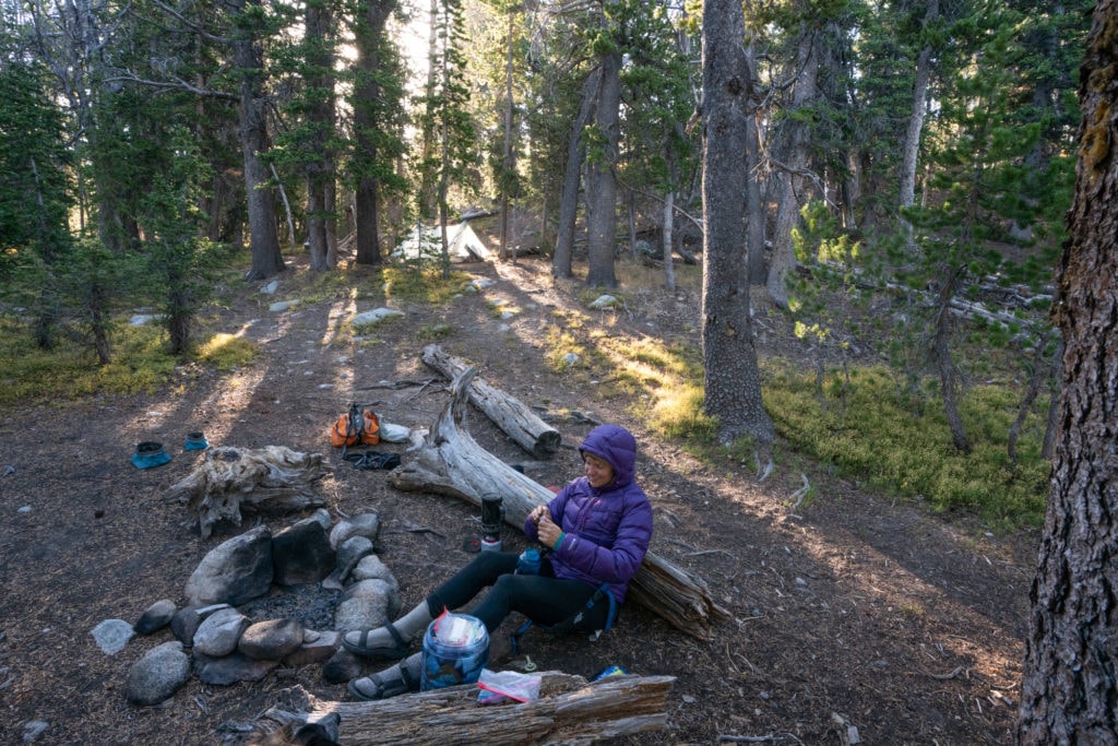 Woman sitting on ground at backcountry campsite with rock fire ring and surrounding forest