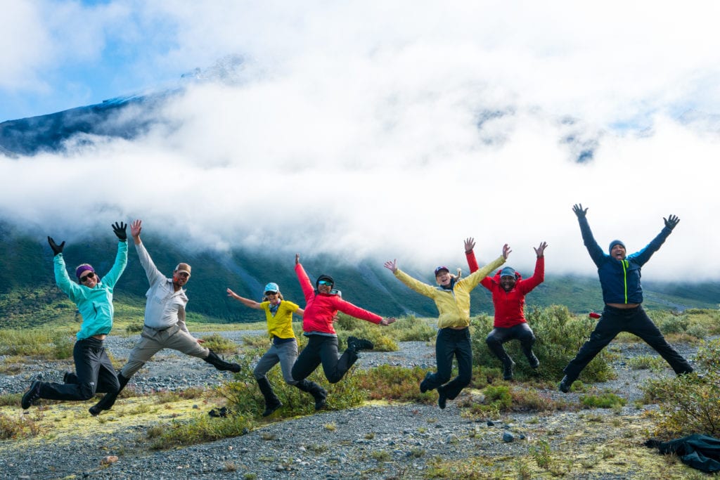 A group of hikers jumping in the air wearing yellow, blue, and red jackets with a low hanging cloud in the background