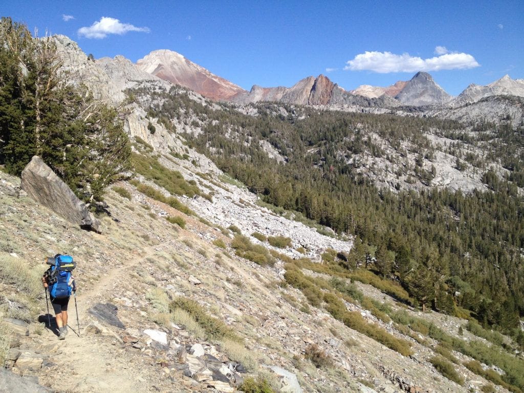 A woman hikes along the John Muir Trail carrying a blue backpacking pack with mountains in the distance